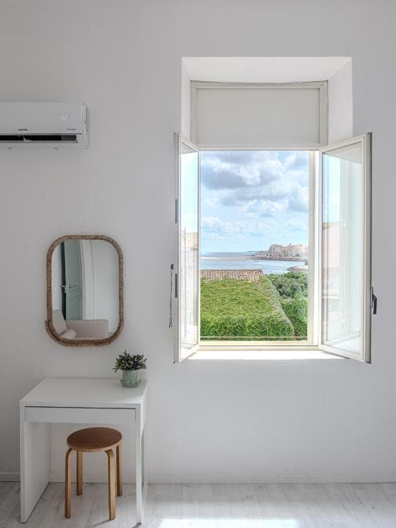 Una ventana del dormitorio abierta al mar: el cielo, el agua y el promontorio en el horizonte, con un espejo y una mesita blanca al lado