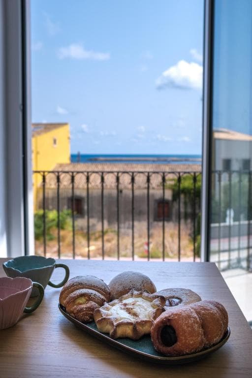 El desayuno en la mesa junto a la ventana abierta: bollería en una bandeja, dos tazas y el mar al fondo tras la barandilla