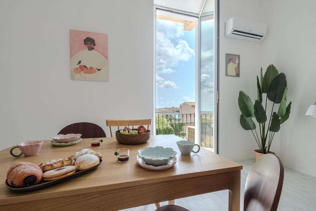 La mesa del comedor puesta para el desayuno ante la puerta del balcón abierta al mar, con la planta y los cuadros en las paredes